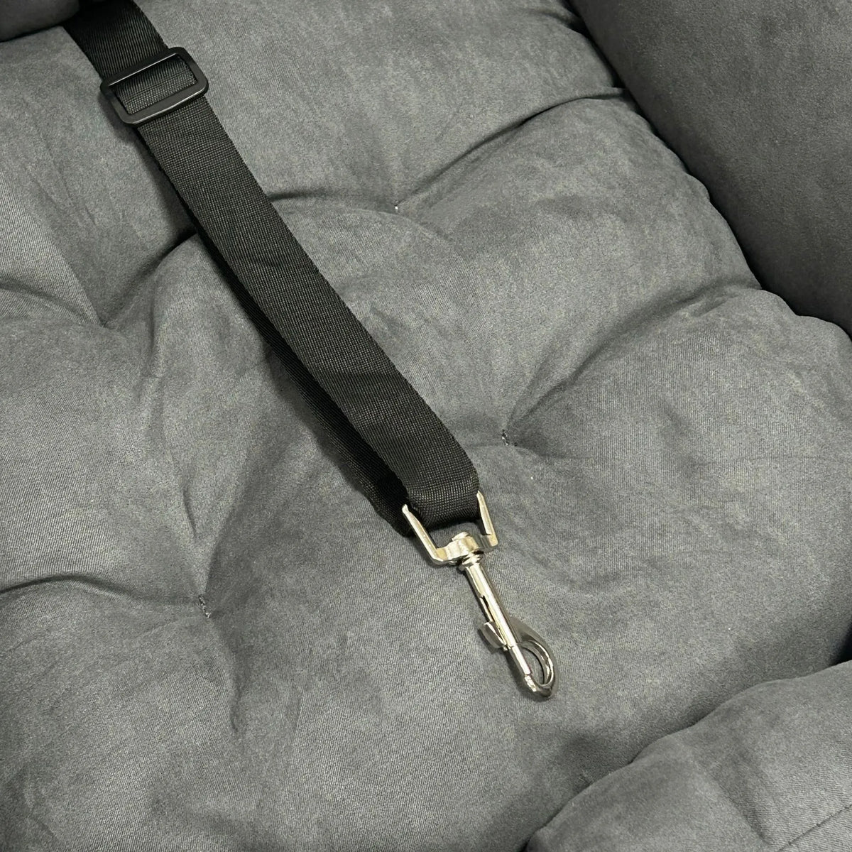 A close-up of a black pet car seat belt with a silver metal clip attached to a gray tufted pet bed.